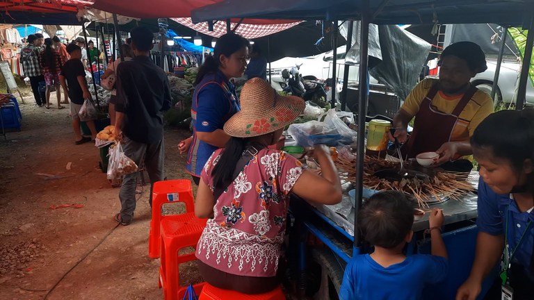 မဲဆောက်မြို့၊ ဟုန်ဖိုင်ရပ်ကွက်အနီးမှ တနင်္ဂနွေစျေးတွင် ရောင်းချနေကြသည့် စျေးဆိုင်များနှင့် လာရောက်စားသောက်နေကြသူများ။ (Photo: RFA)