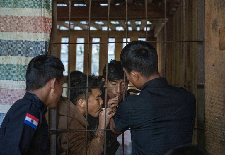 ရှမ်းပြည်နယ်တောင်ပိုင်းရှိ ကရင်နီပြည်ရဲဌာန အမှတ် (၈) စခန်းတွင် ထိန်းသိမ်းထားသူနှစ်ဦးကို တာဝန်ကျရဲအရာရှိတစ်ဦးက ဆေးလိပ်မီးညှိပေးနေသည်ကို ၂၀၂၄ ခုနှစ် နိုဝင်ဘာ ၄ ရက်နေ့က တွေ့ရစဉ်။ (Gemunu Amarasinghe/RFA)